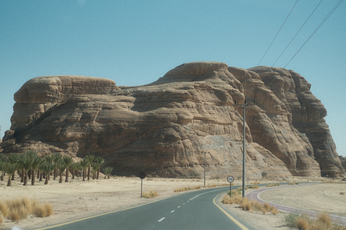 Massive sandstone rock formation with a desert road and palm trees in AlUla, Saudi Arabia