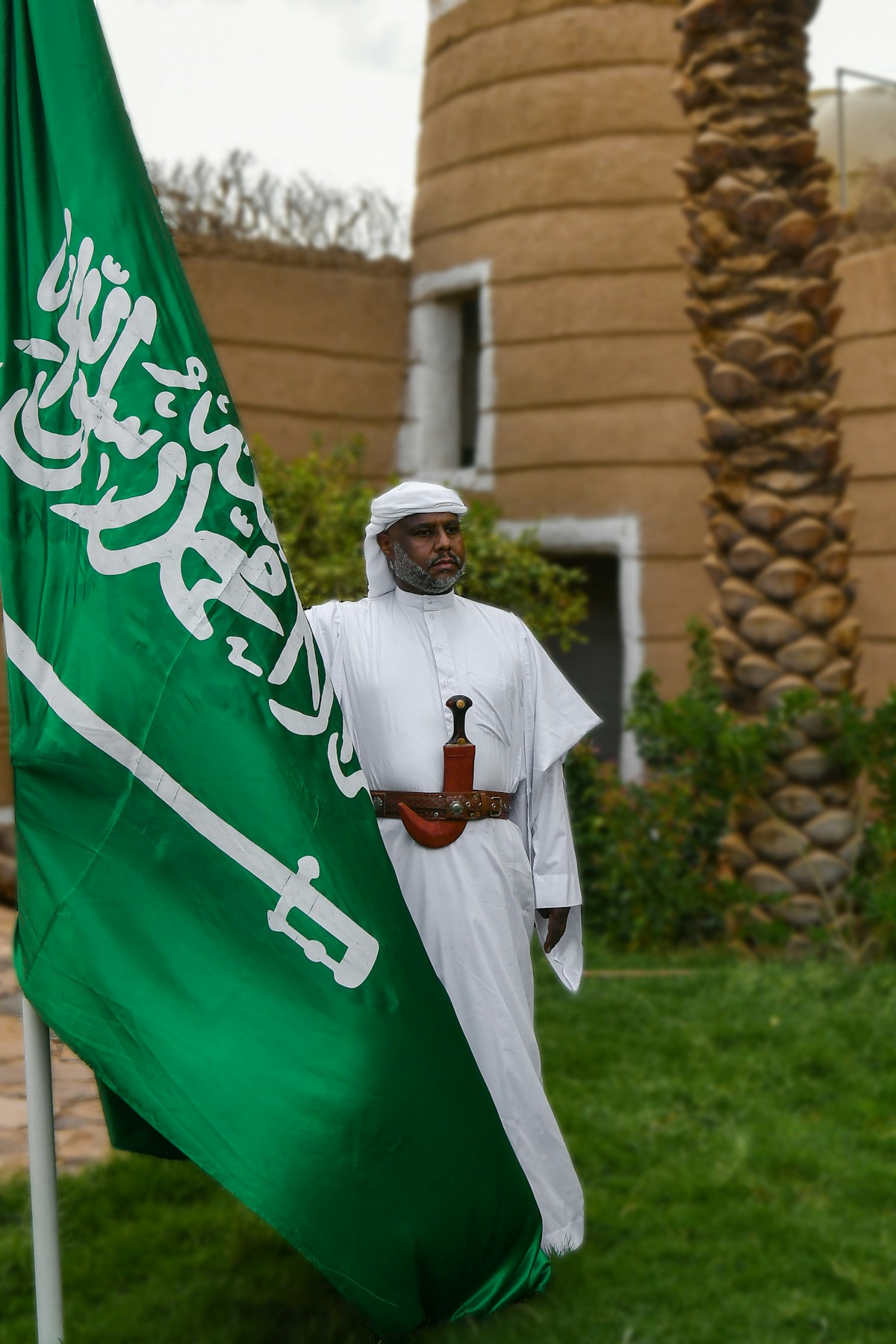 Saudi man in traditional thobe holding the national flag near a historic mud-brick tower in southern Saudi Arabia