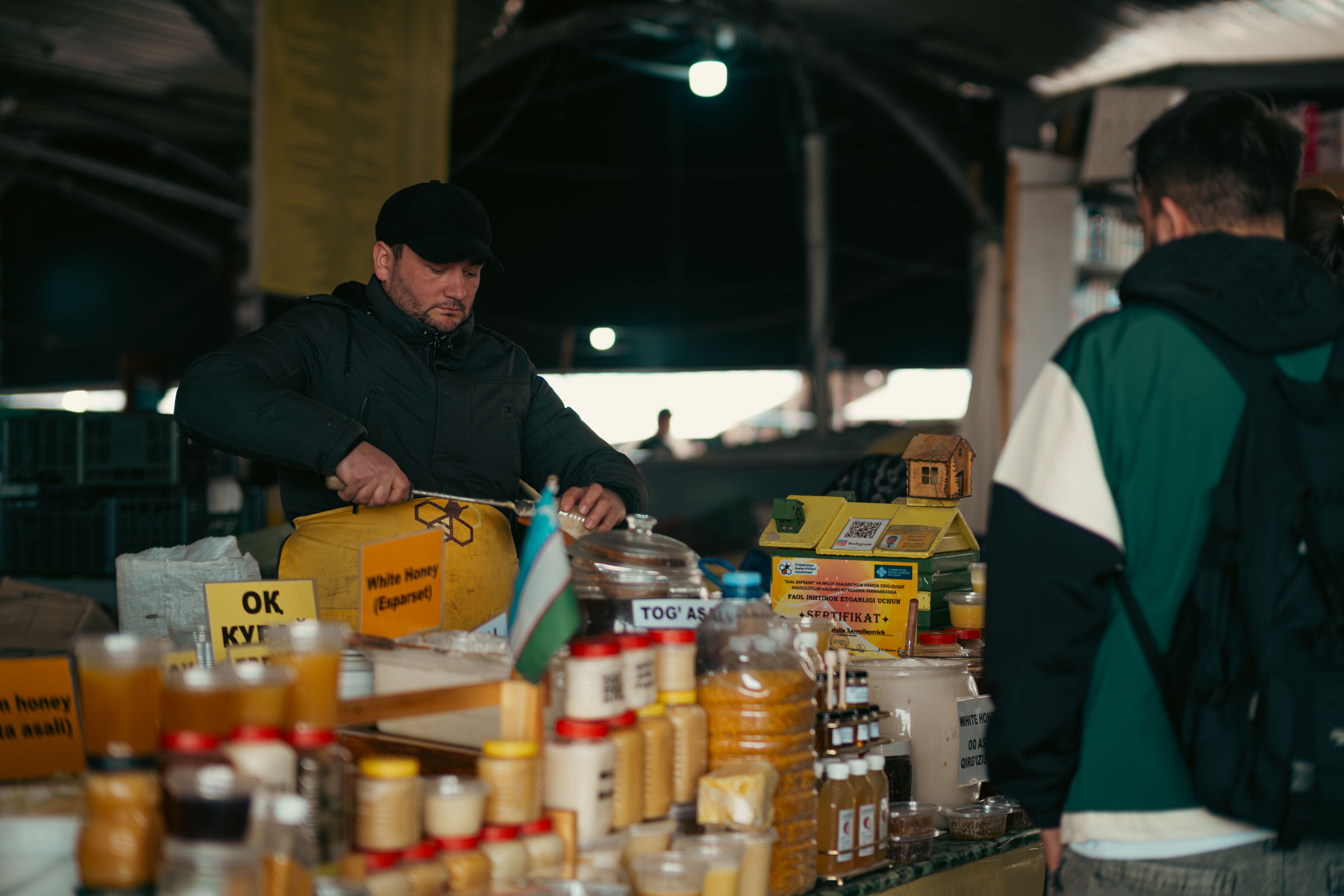 Vendor tending rows of honey jars and bottles at an indoor market stall with warm atmospheric lighting