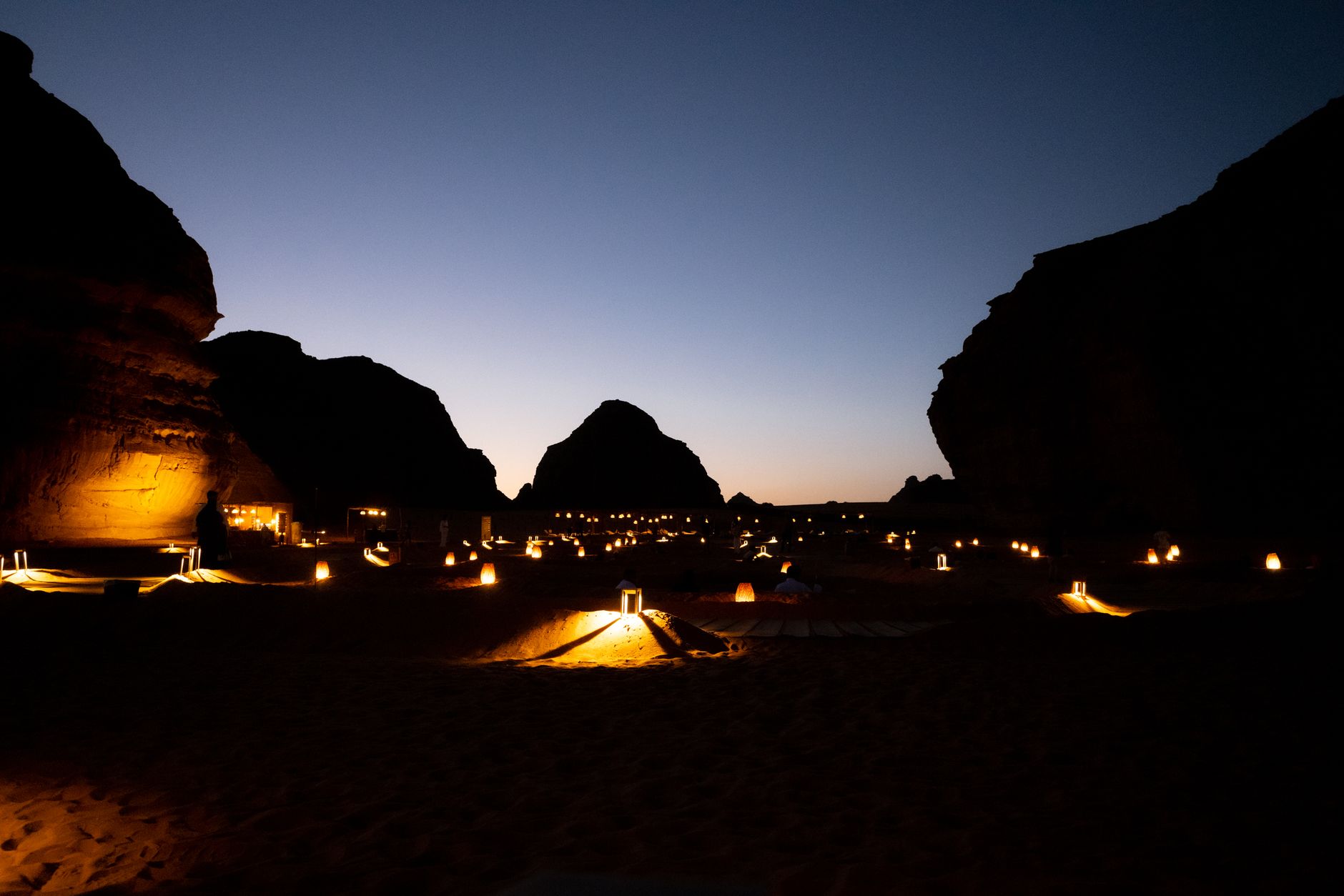 Desert camp at night in AlUla lit by lanterns with dramatic rock formations silhouetted against a deep blue sky